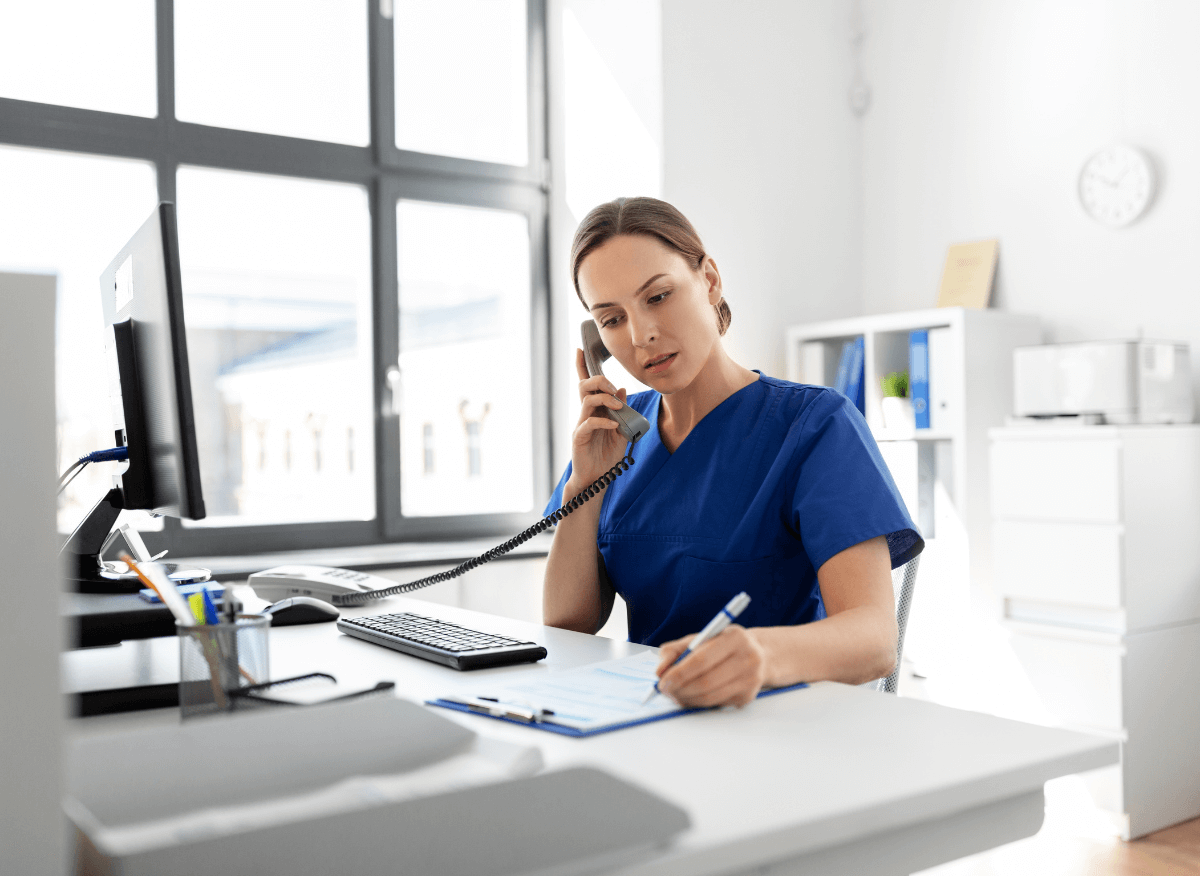 A woman in blue scrubs is on the phone at a desk, writing on a clipboard. A woman in blue scrubs is on the phone at a desk, writing on a clipboard.